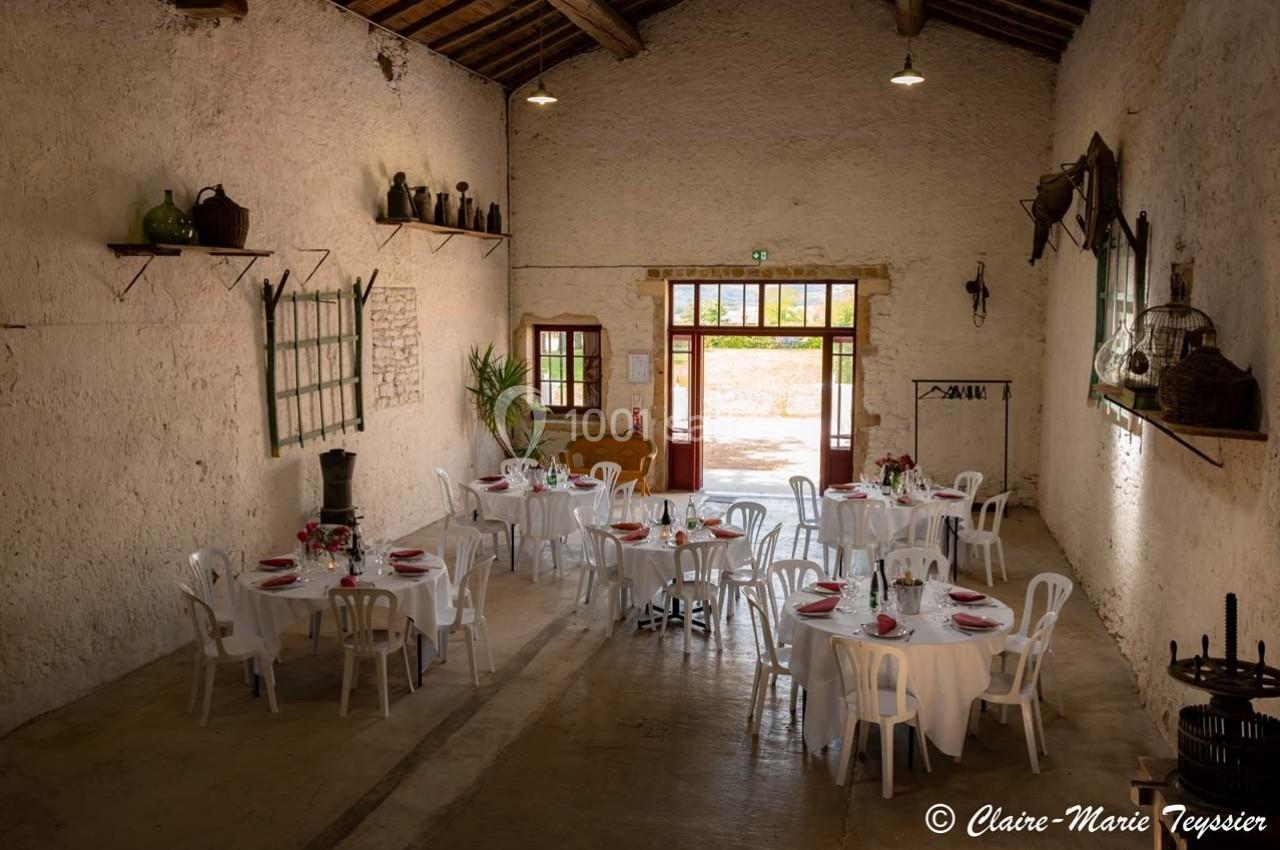 Salle rustique avec murs en pierre, tables rondes dressées avec nappes blanches et chaises, éclairée par des portes vitrées.