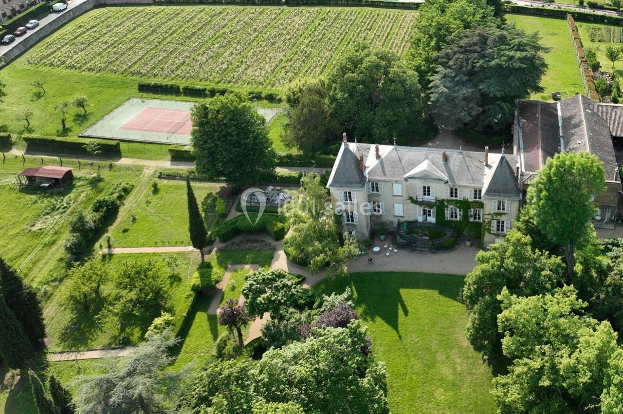 Vue aérienne d'une maison entourée de jardins, d'un terrain de tennis et de vignes en arrière-plan.