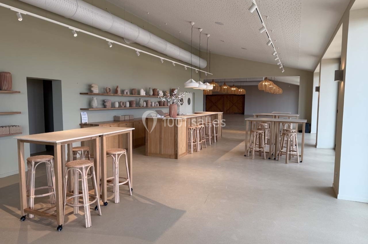 Salle lumineuse avec tables hautes en bois, tabourets, étagères décorées de céramiques et suspensions en osier.
