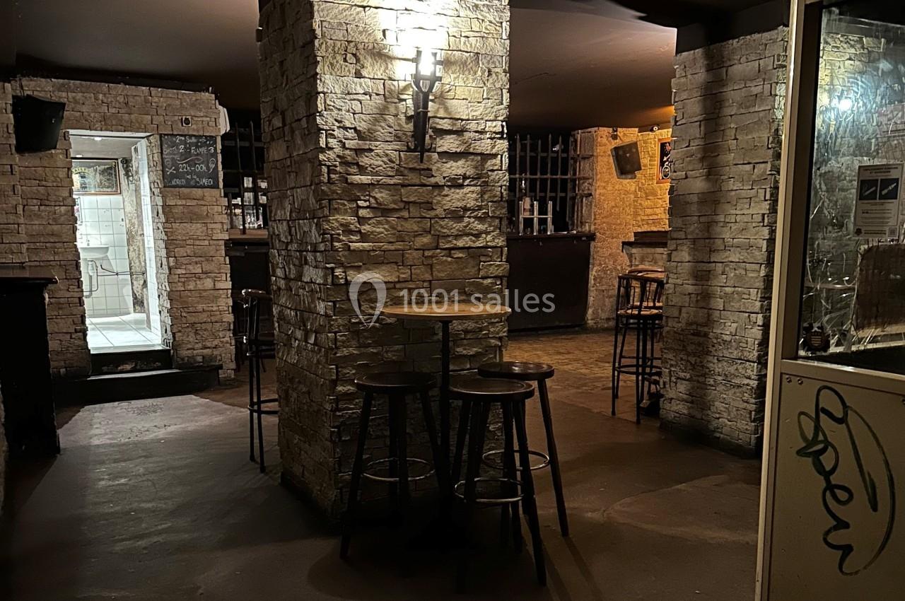 Intérieur d'un bar sombre avec des murs en pierre, des tabourets autour d'une table et un éclairage tamisé.