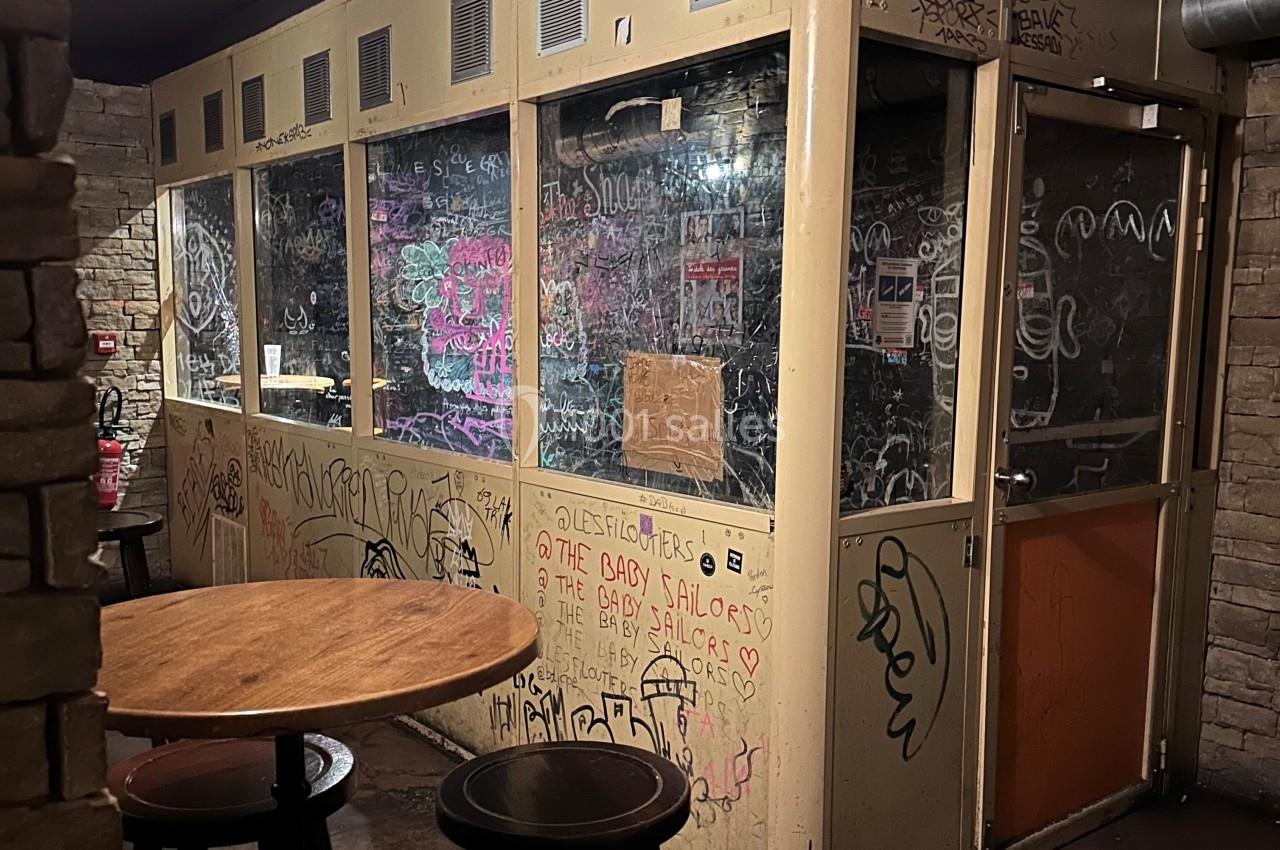 Cabine vitrée dans un espace intérieur, recouverte de graffitis et tags, avec une table ronde au premier plan.