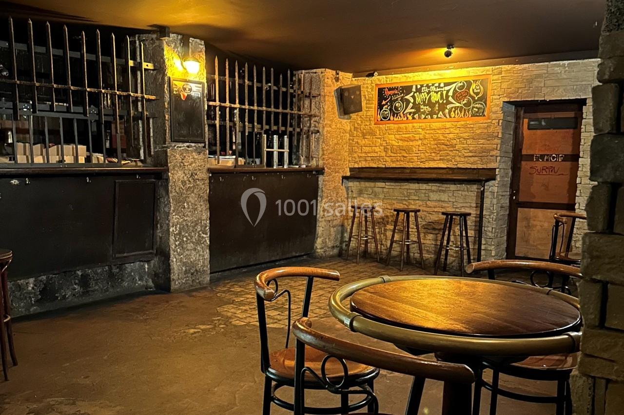 Salle de bar sombre avec tables en bois, tabourets hauts, mur en briques et tableau noir décoratif.