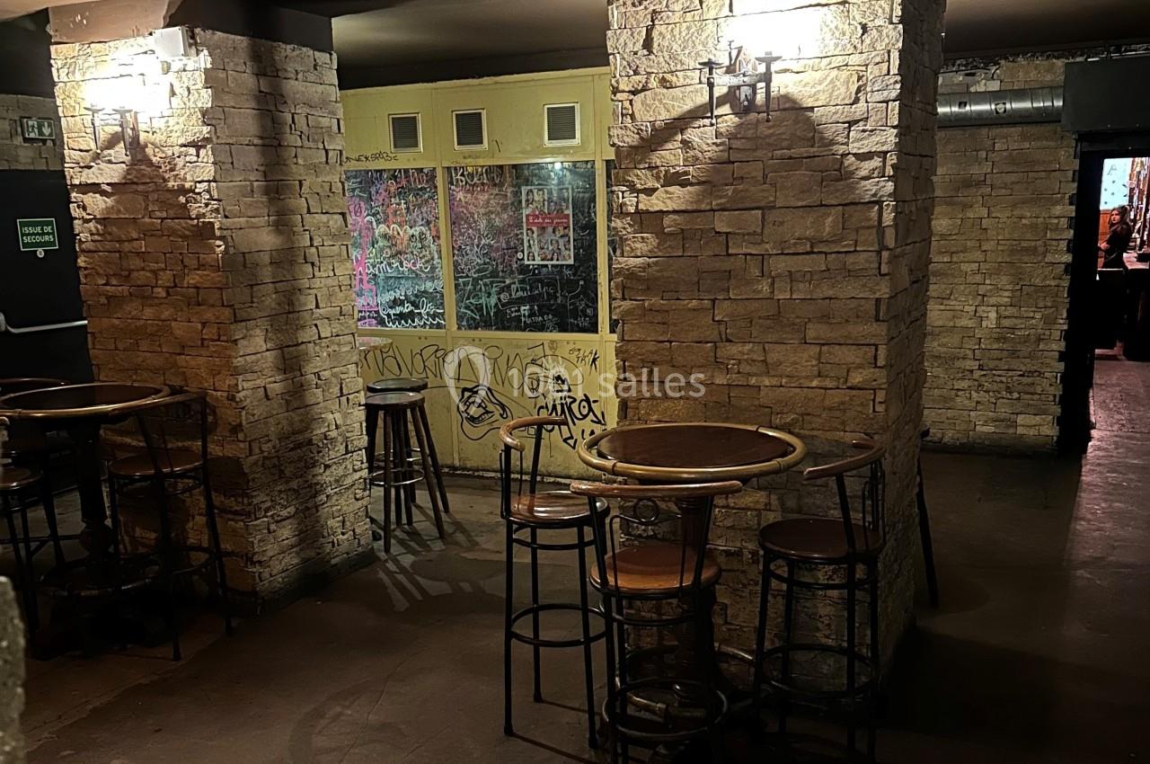 Salle avec murs en pierre, tables hautes en bois et graffitis sur une vitre au fond, éclairée par des appliques murales.