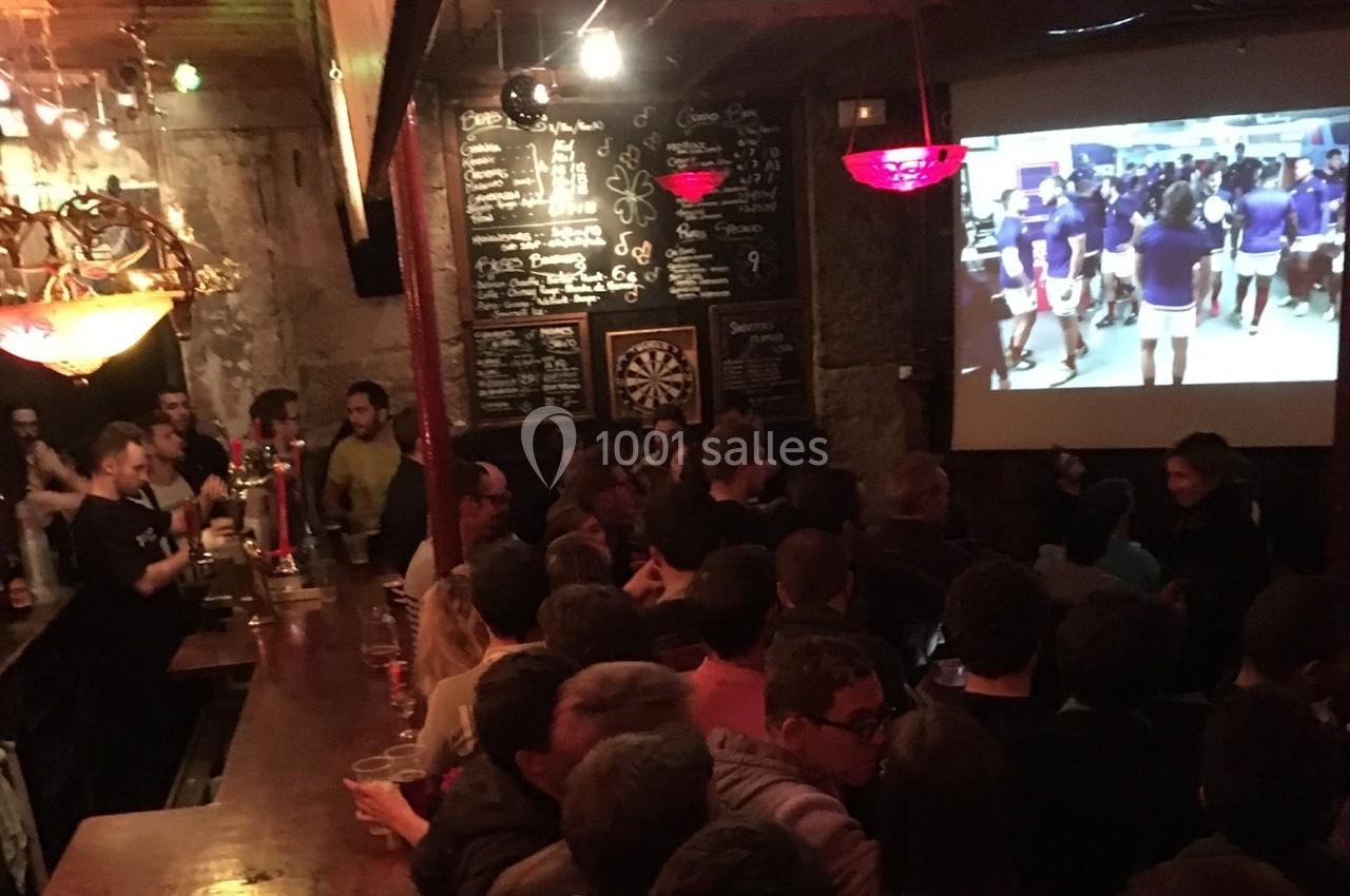 Bar bondé avec des clients regardant un écran diffusant un événement sportif, ambiance animée et conviviale.