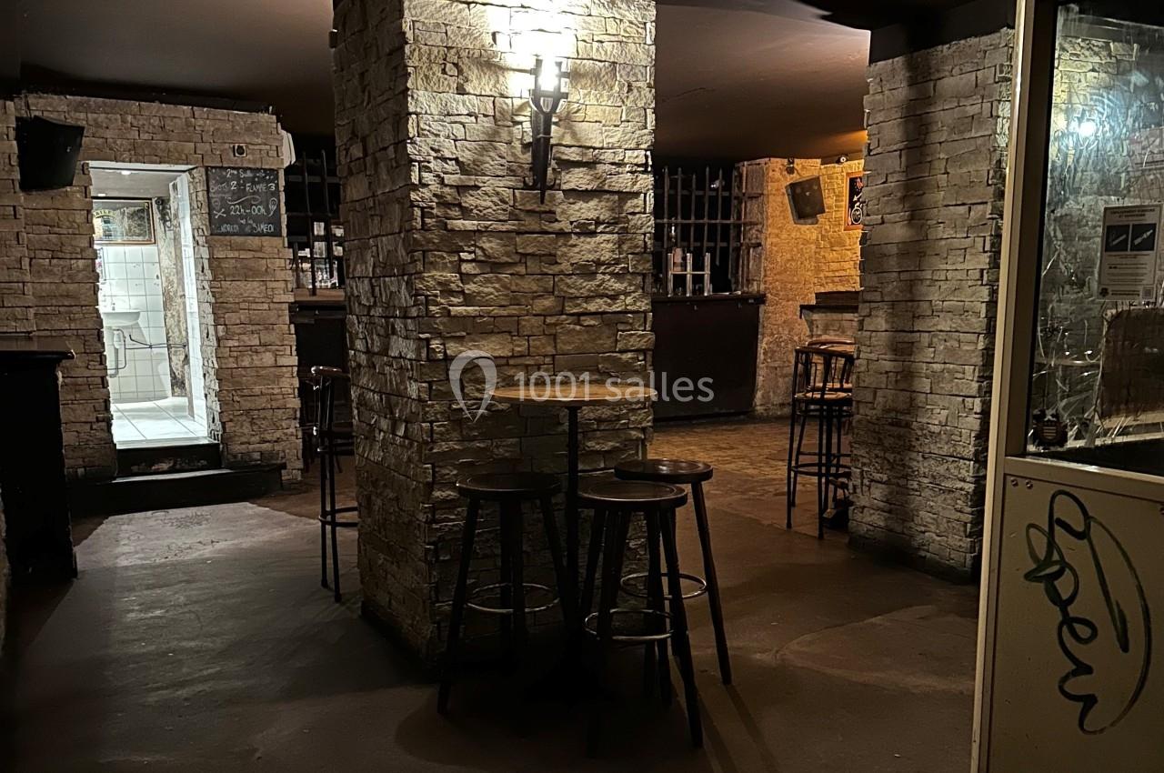 Salle sombre avec murs en pierre, tables hautes et tabourets en bois, éclairée par une applique murale.