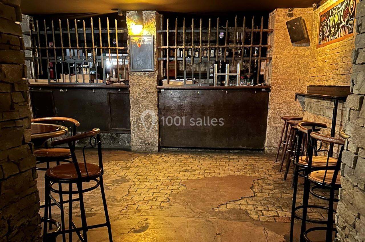Intérieur d'un bar avec comptoir en bois, tabourets hauts et murs en pierre dans une ambiance tamisée.