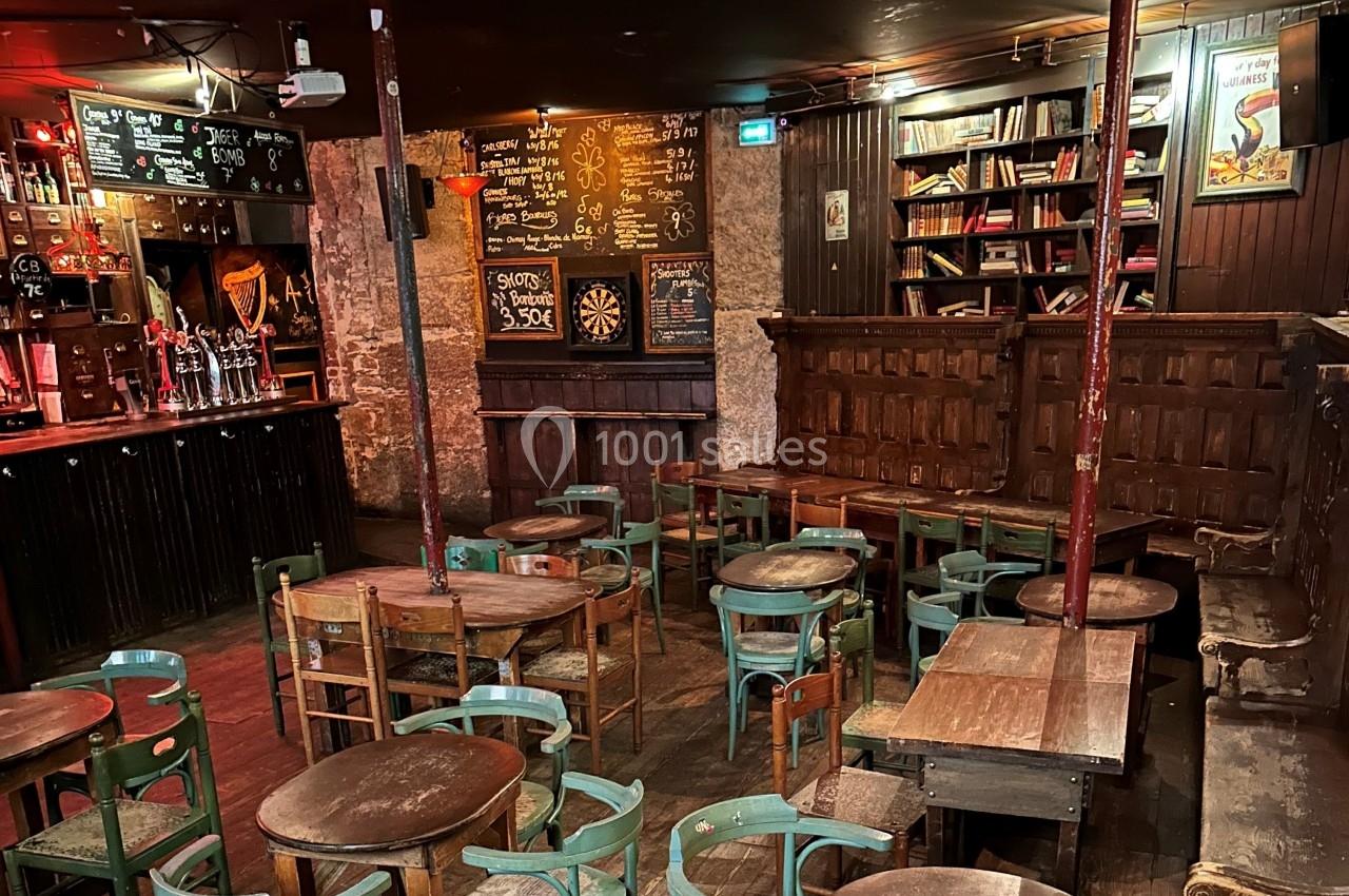Intérieur d'un bar avec tables et chaises en bois, tableau noir avec menus, étagères de livres et éclairage tamisé.