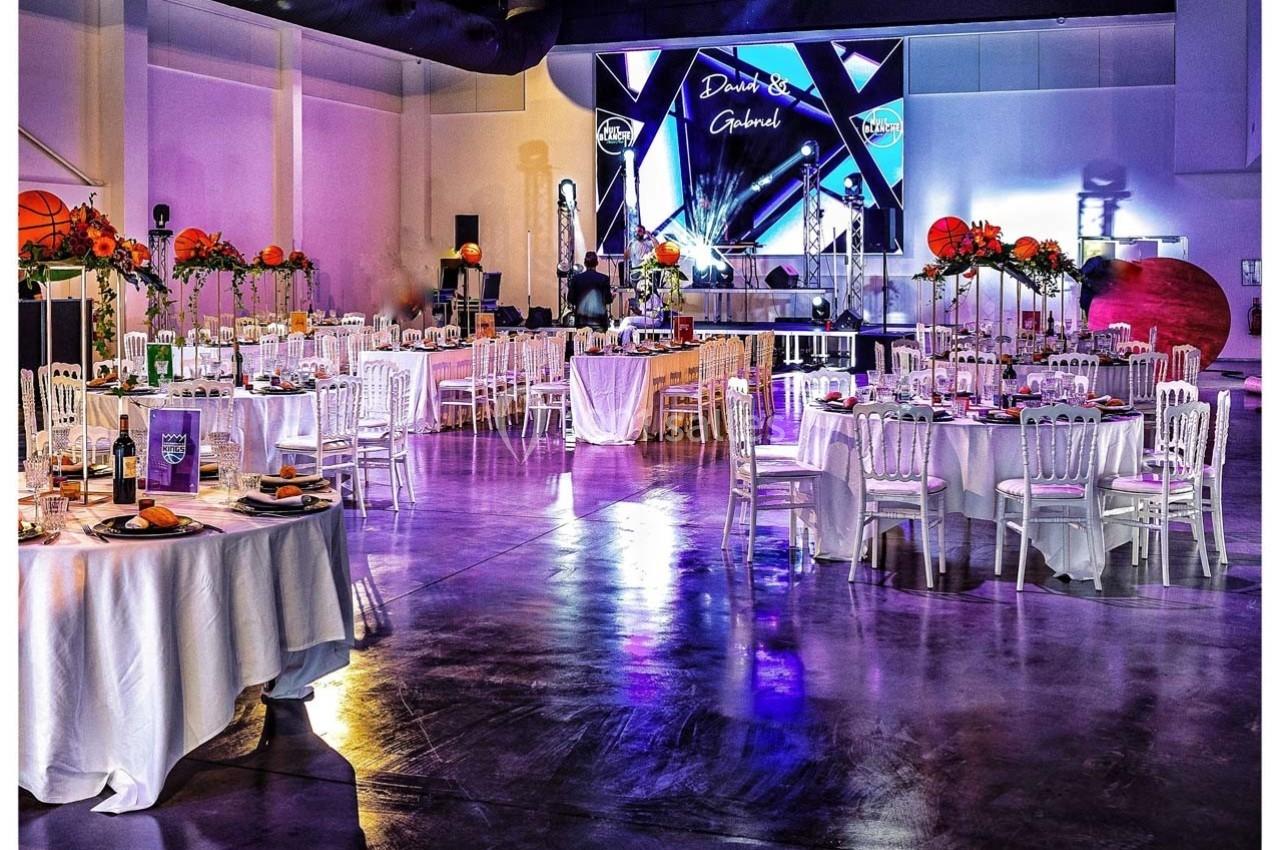 Salle de réception décorée avec des tables dressées, des chaises blanches et une scène illuminée en arrière-plan.