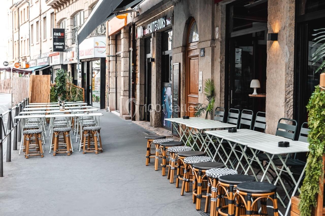 Terrasse de café avec tables et chaises alignées sur un trottoir, bordée de plantes et devantures de magasins.
