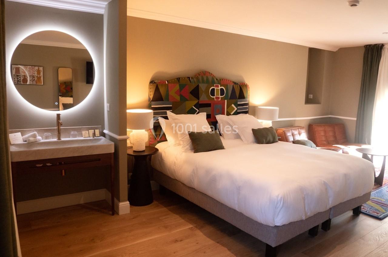 Chambre d'hôtel lumineuse avec lit double, tête de lit colorée, miroir rond éclairé et coin salon.