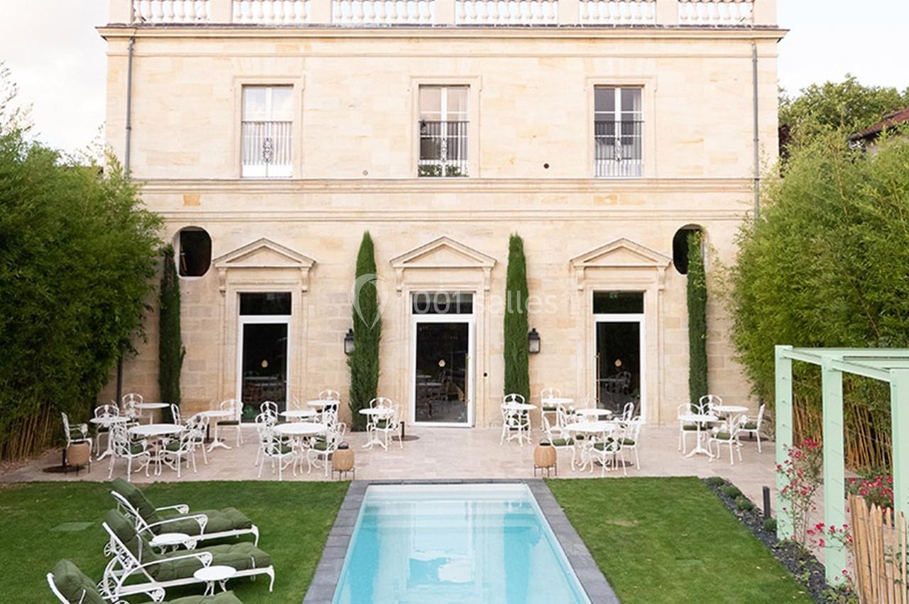 Façade d'un bâtiment en pierre avec terrasse aménagée, tables, chaises blanches et piscine au premier plan.