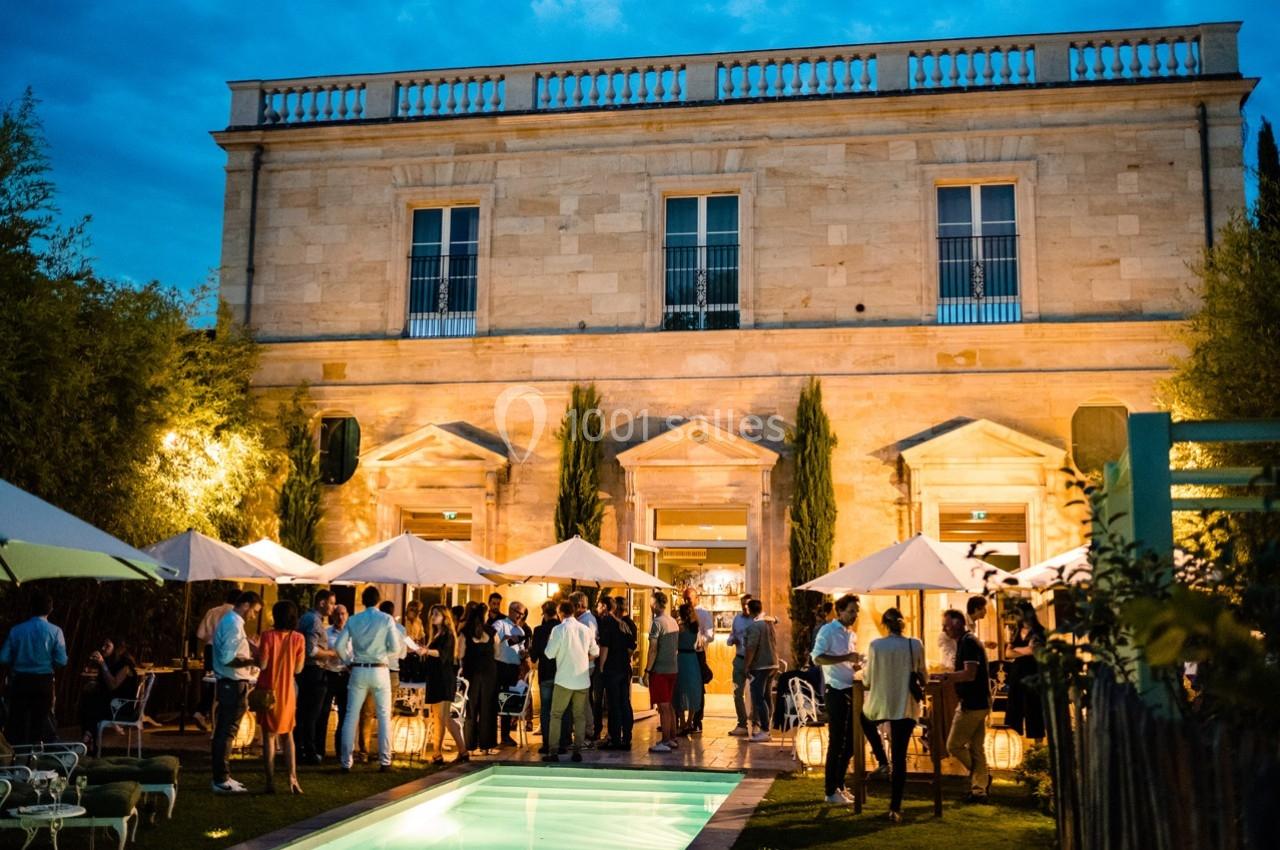 Groupe de personnes réunies en soirée dans un jardin éclairé, devant une grande bâtisse en pierre.