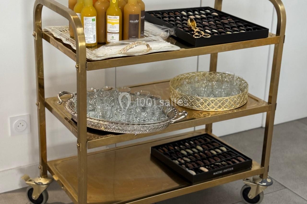 Chariot doré à trois étagères contenant des bouteilles de jus, des verres, et des plateaux de chocolats.