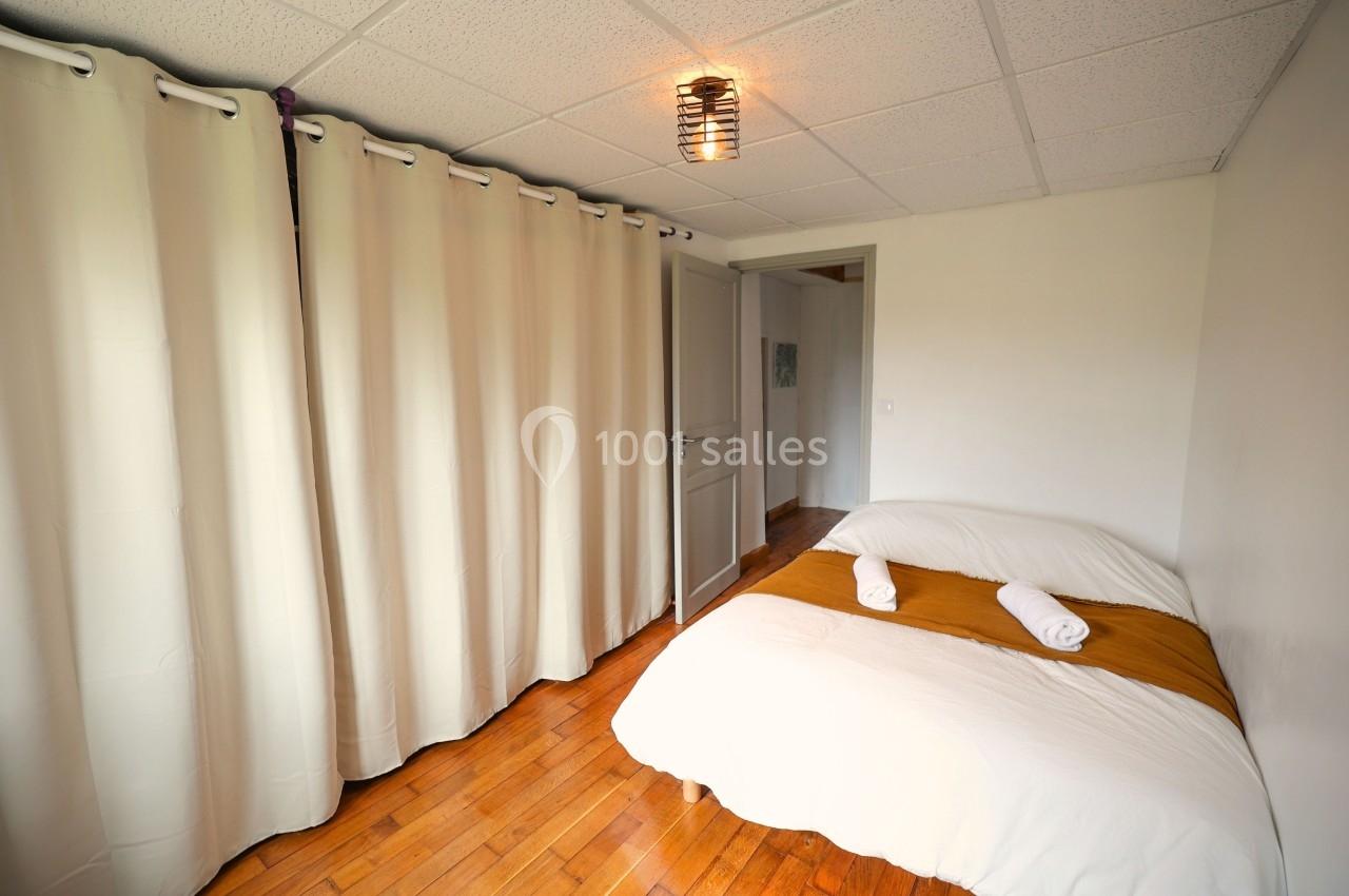 Chambre lumineuse avec un lit double, draps blancs, plaid ocre, rideaux beiges et parquet en bois.