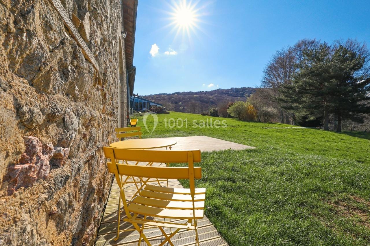 Chaises et tables jaunes sur une terrasse en bois, près d'un mur en pierre, sous un ciel ensoleillé avec vue sur la campagne.