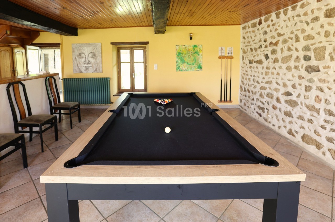 Table de billard noir dans une pièce lumineuse avec murs en pierre, chaises en bois et décorations murales.