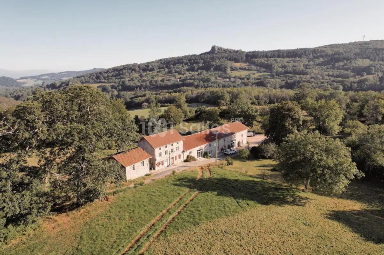 Vue aérienne d'un ensemble de bâtiments en pierre avec toits rouges, entouré de verdure et de collines boisées.