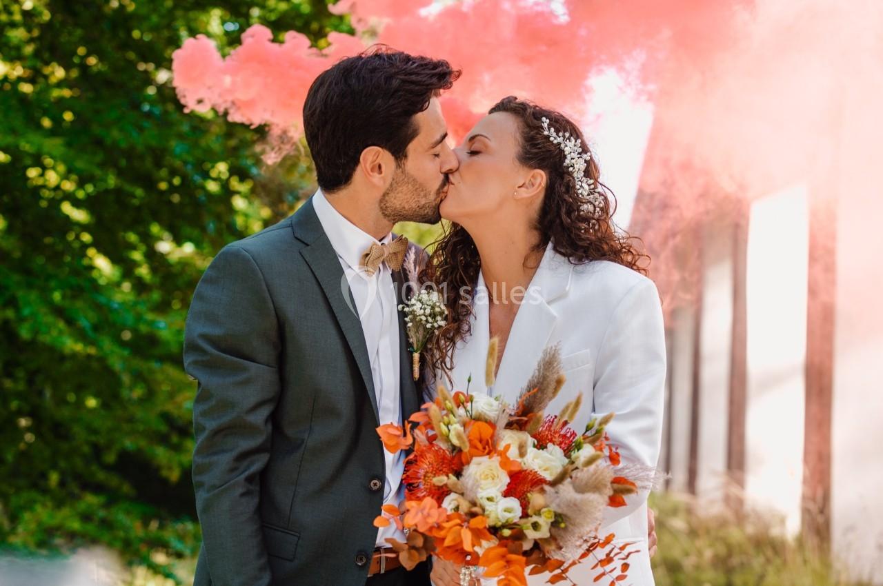 Un couple s'embrasse lors d'un mariage, tenant un bouquet coloré, avec de la fumée rose en arrière-plan.