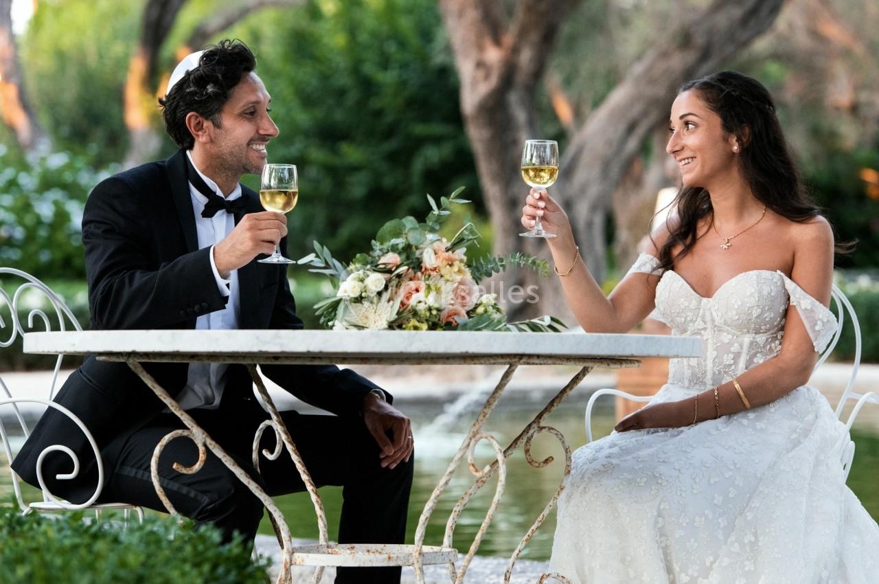 Un couple en tenue de mariage trinque avec des verres de vin blanc, assis à une table en extérieur.