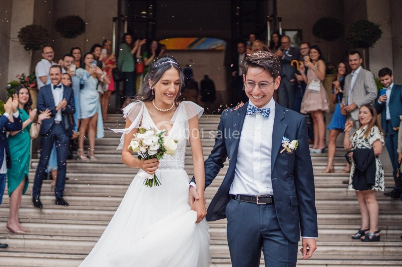 Un couple souriant sort d'un bâtiment après leur mariage, entouré d'invités applaudissant sur les marches.
