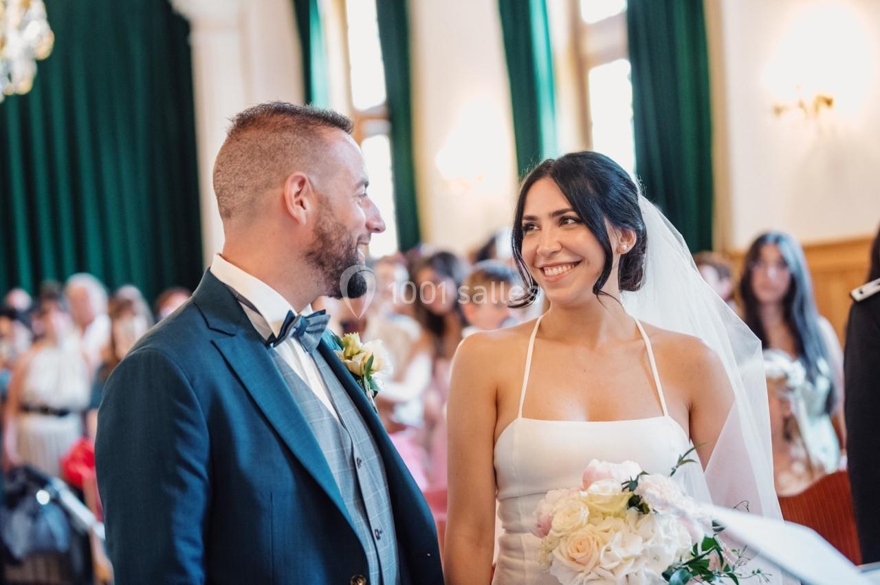 Un couple souriant lors de leur cérémonie de mariage dans une salle ornée de rideaux verts.