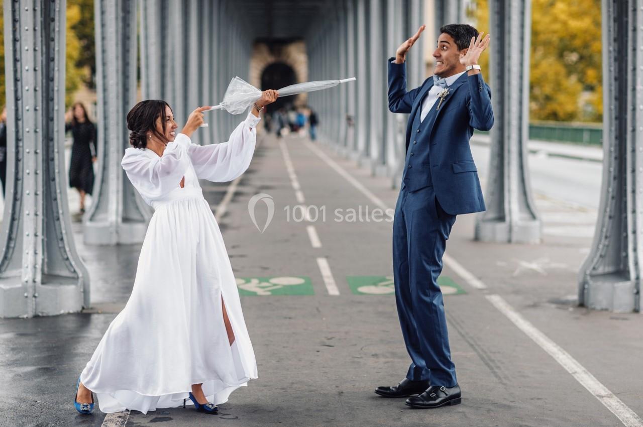 Une femme en robe blanche joue avec un parapluie face à un homme en costume sur un pont métallique.