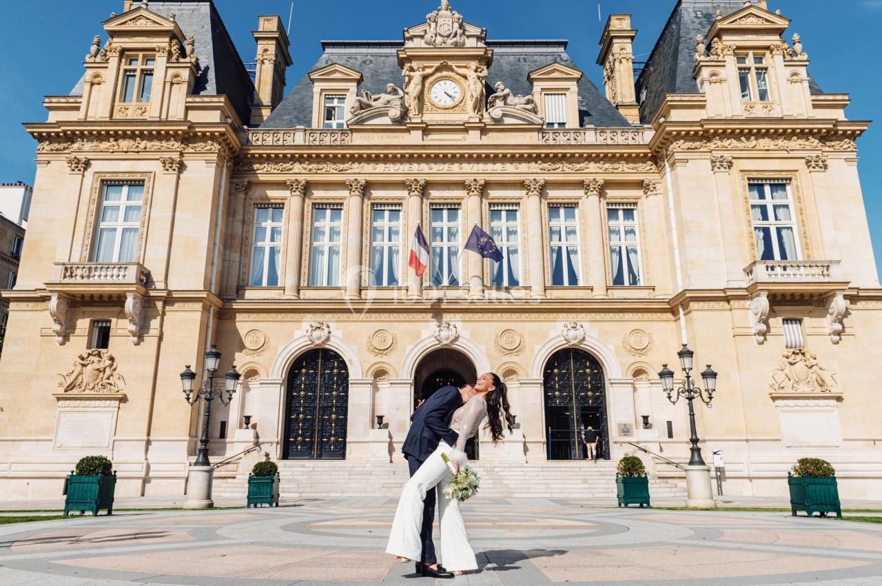 Un couple en tenue de mariage s'embrasse devant un bâtiment historique avec des drapeaux français et européen.