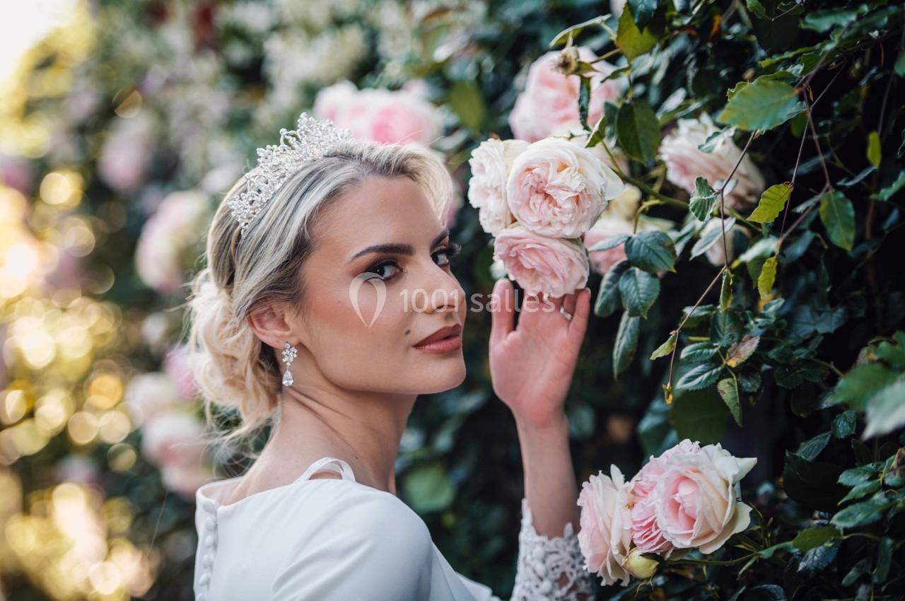 Femme en robe blanche et diadème, posant près d'un mur couvert de roses roses dans un jardin ensoleillé.
