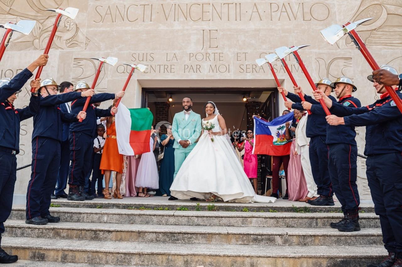 Un couple de mariés sort d'un bâtiment, entouré de pompiers formant une haie d'honneur avec des haches.