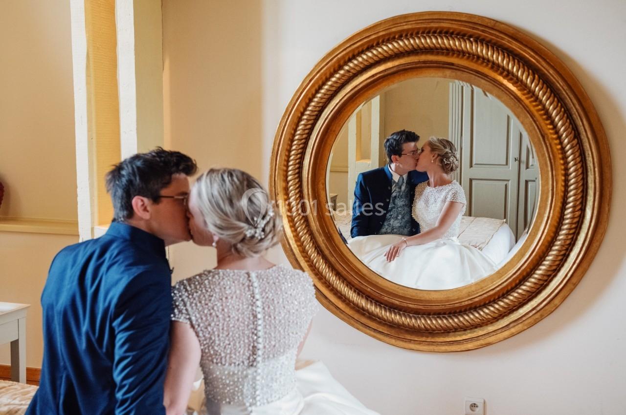 Un couple en tenue de mariage s'embrasse, leur reflet visible dans un miroir rond encadré de doré.
