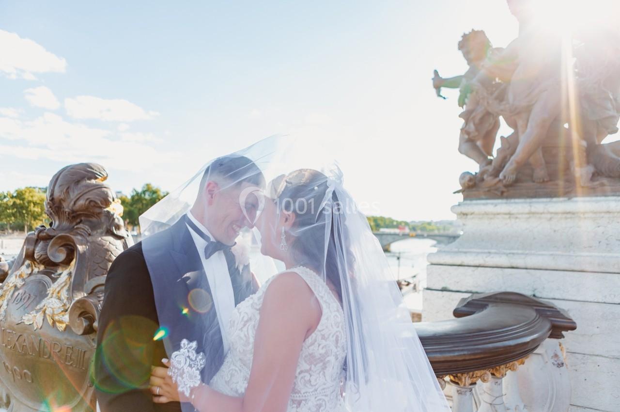 Un couple de mariés sourit sous un voile, près d'une sculpture en plein air par une journée ensoleillée.