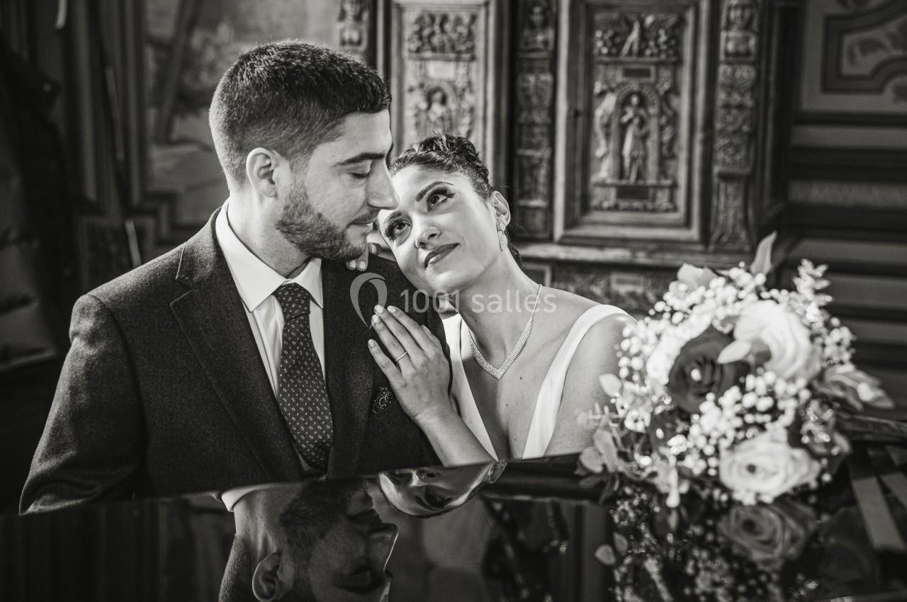 Un couple habillé élégamment partage un moment tendre près d'un piano, avec un bouquet de fleurs en avant-plan.