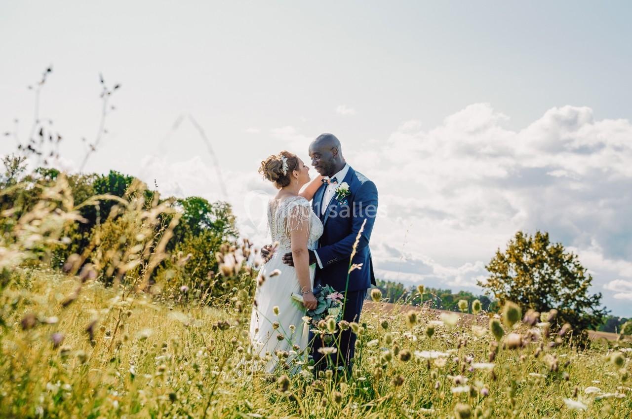 Un couple en tenue de mariage se tient dans un champ fleuri sous un ciel partiellement nuageux.