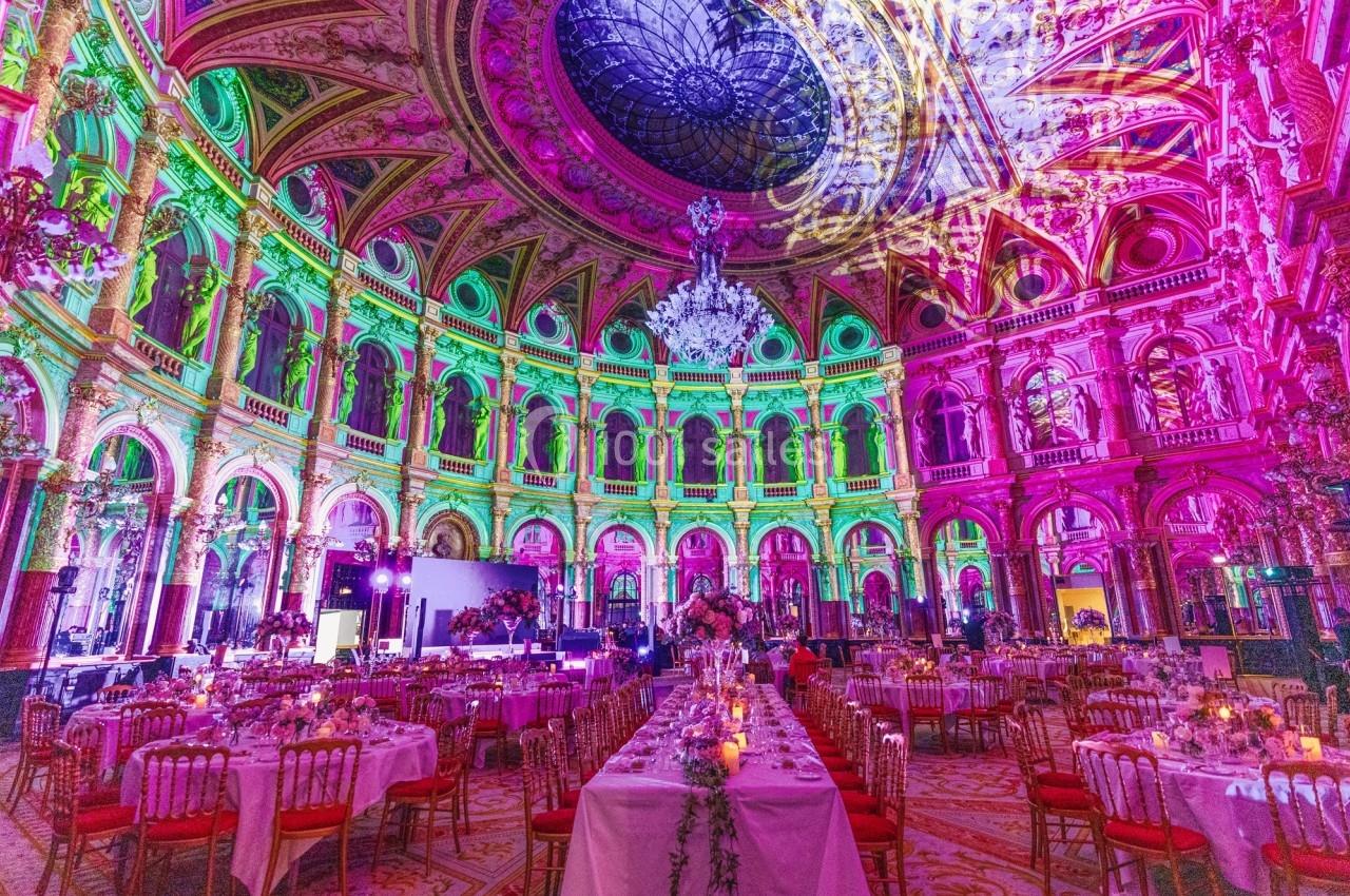 Salle de réception luxueuse avec plafond orné, éclairage coloré et tables dressées pour un événement.