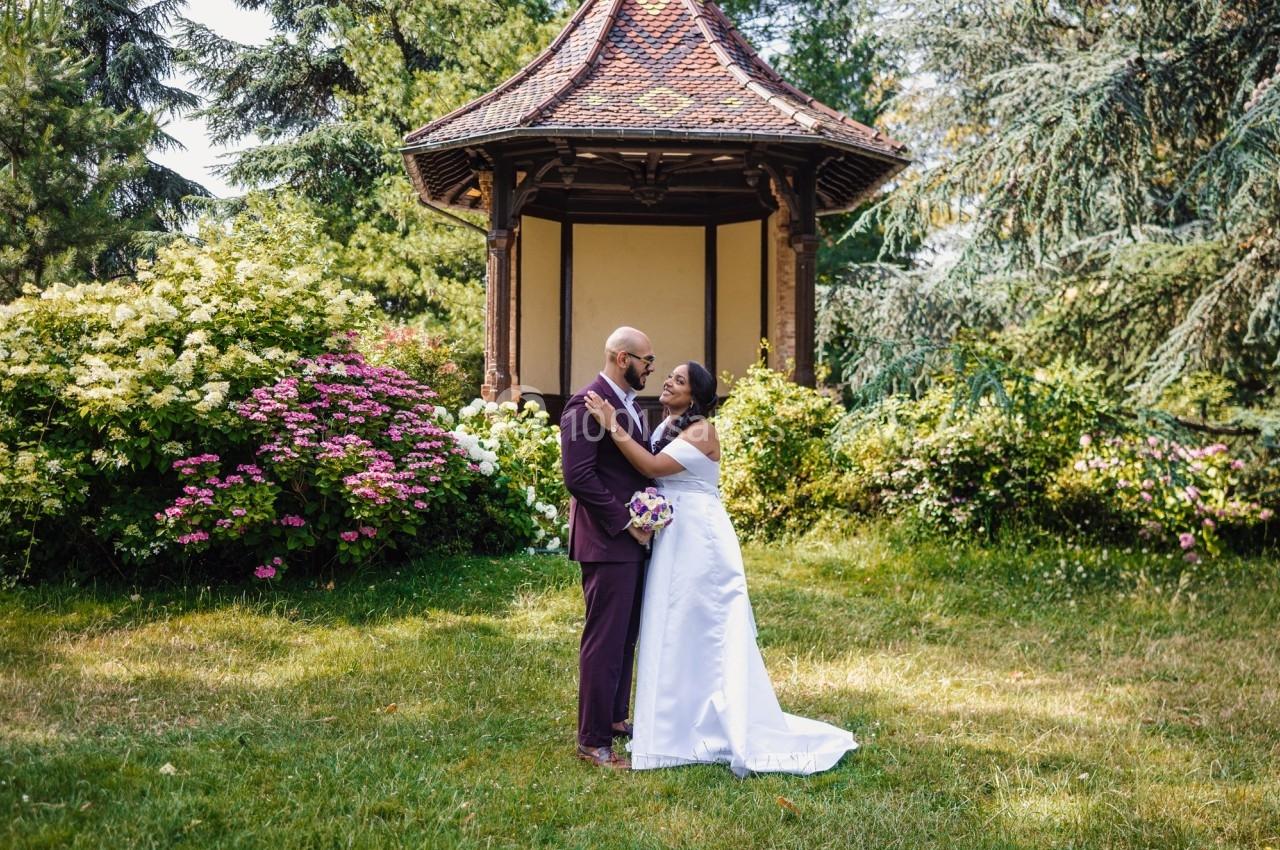 Un couple en tenue de mariage pose dans un jardin verdoyant devant un pavillon en bois.