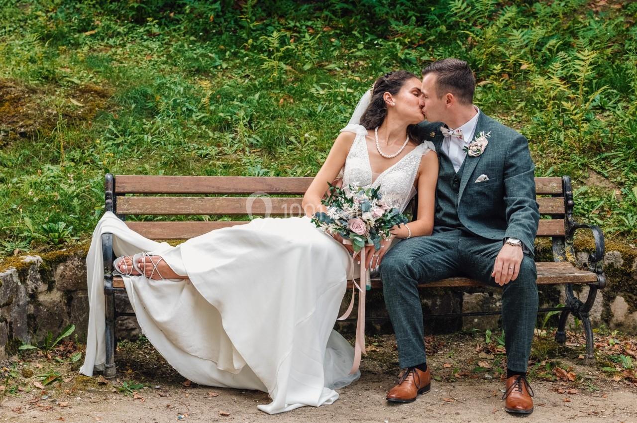 Un couple de mariés s'embrasse sur un banc en bois dans un cadre naturel verdoyant.