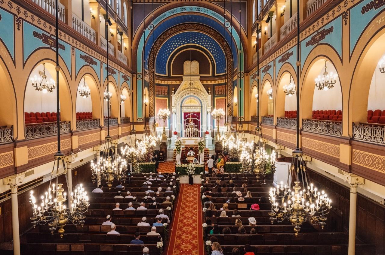 Intérieur d'une synagogue avec des fidèles assis, un autel central décoré et des lustres suspendus.
