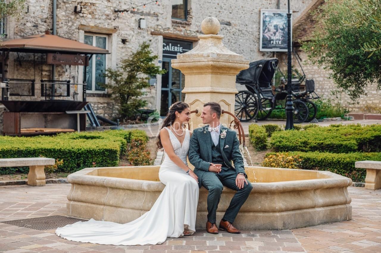 Un couple en tenue de mariage est assis sur le rebord d'une fontaine en pierre dans une cour pavée.