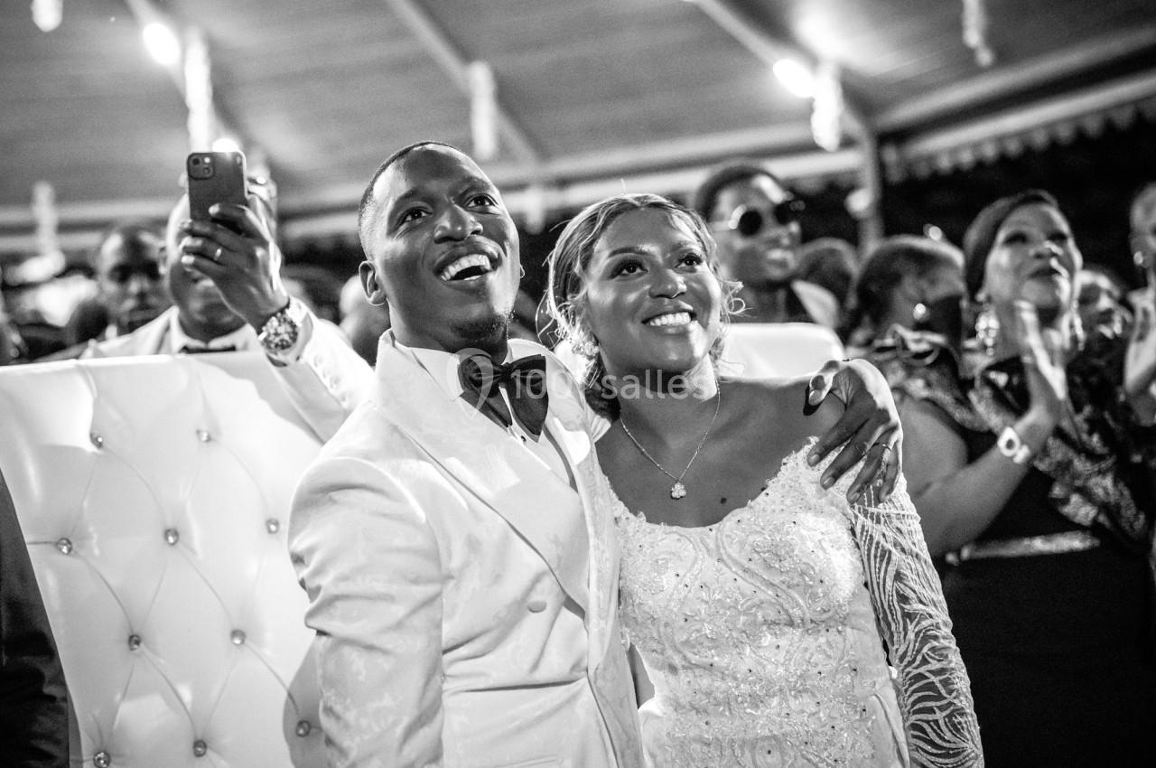 Un couple en tenue de mariage sourit, entouré d'invités dans un cadre festif en noir et blanc.