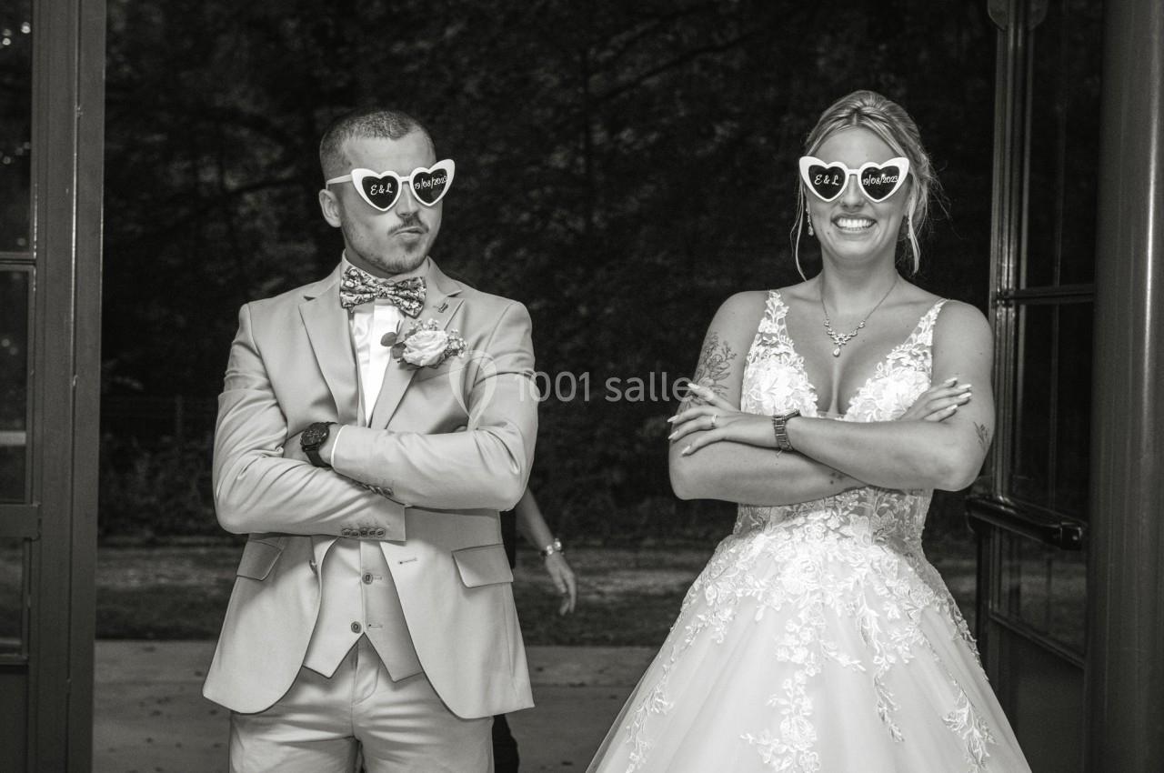 Un couple en tenue de mariage pose avec des lunettes fantaisie en forme de cœur, bras croisés et sourires amusés.