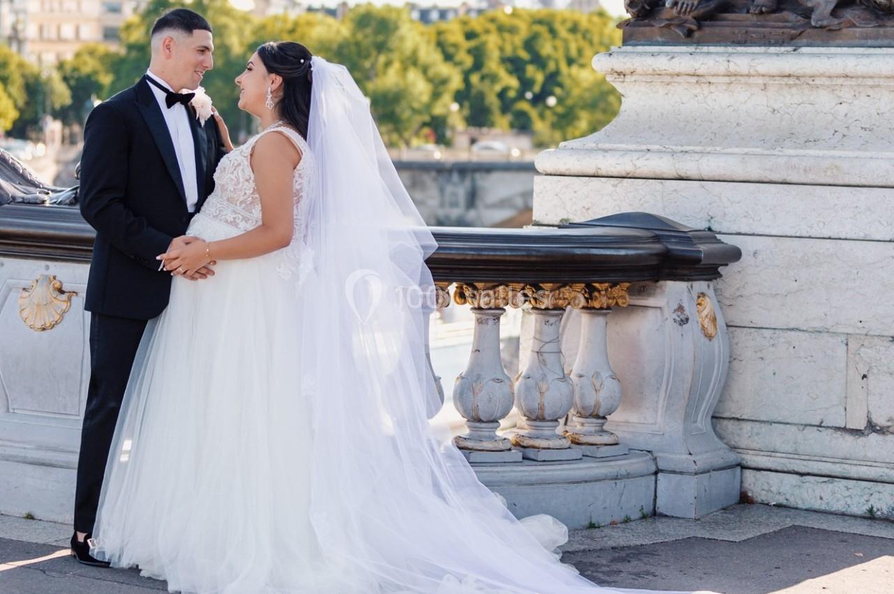 Un couple en tenue de mariage pose sur un pont orné, avec des arbres et un ciel dégagé en arrière-plan.