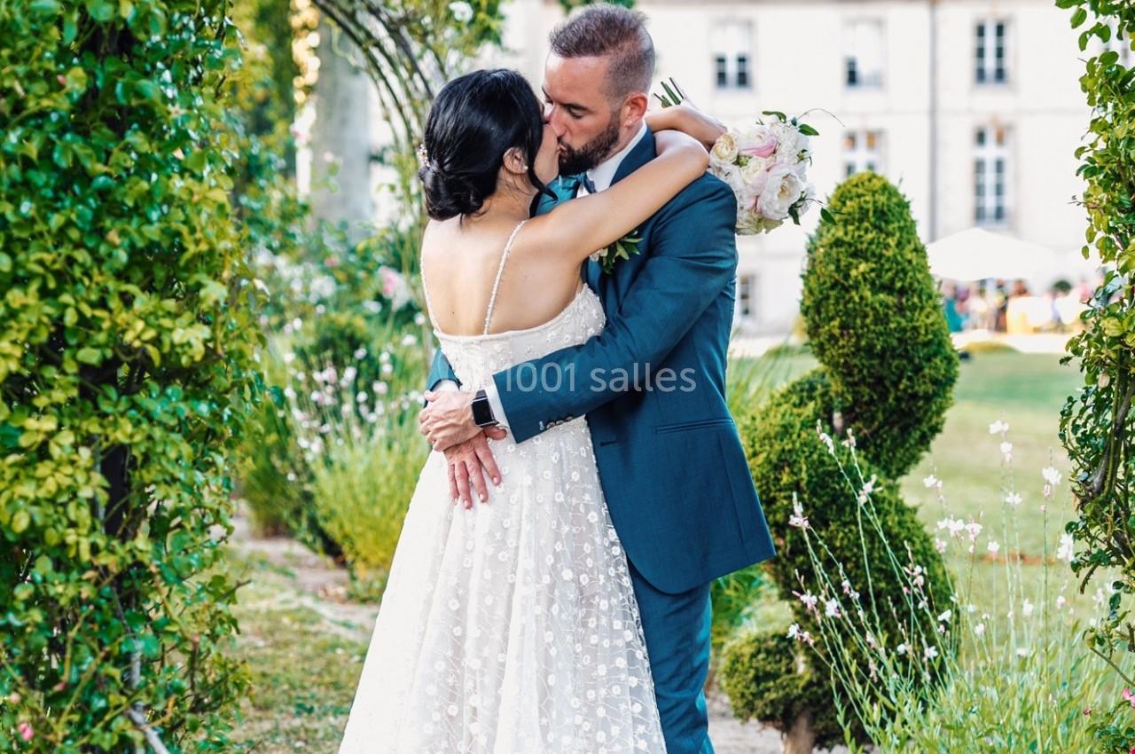 Un couple en tenue de mariage s'embrasse dans un jardin verdoyant devant un bâtiment élégant.