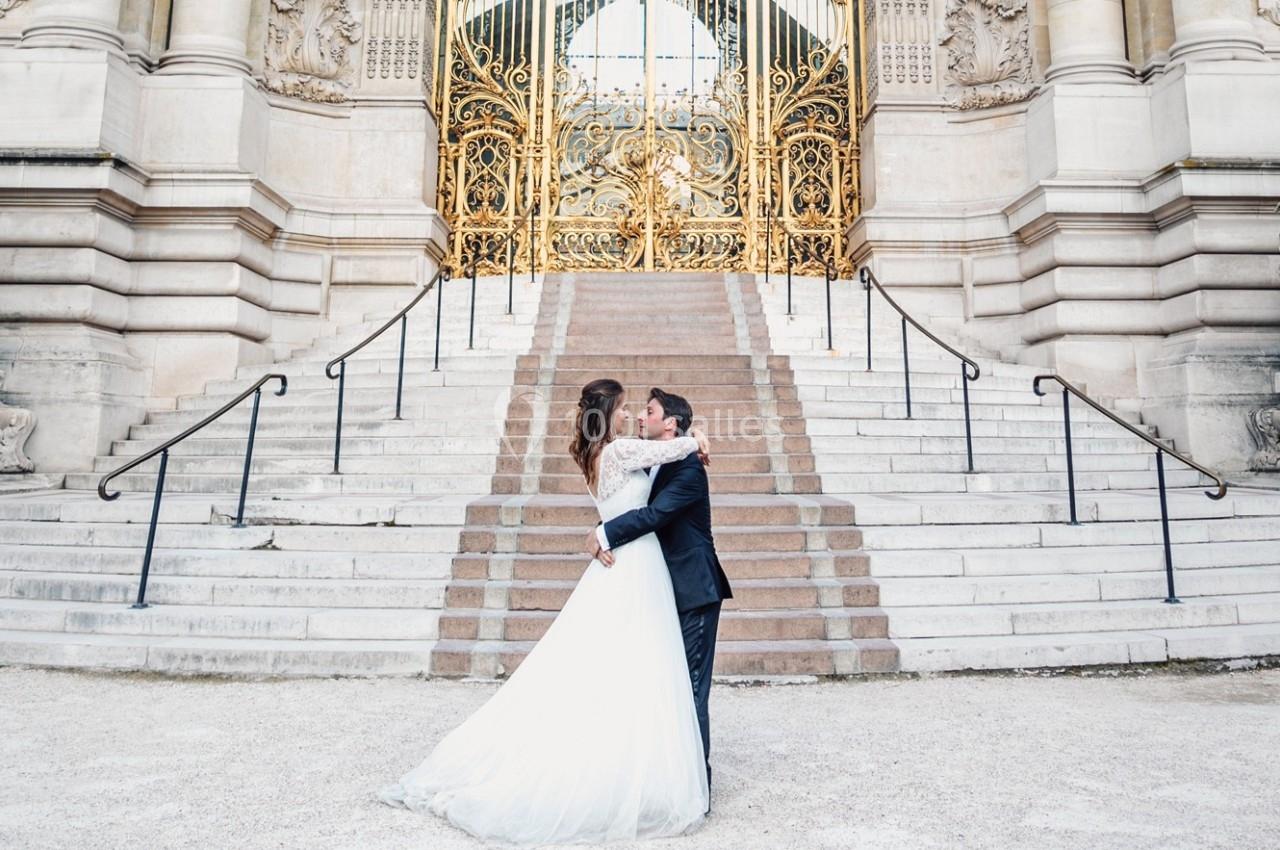 Un couple en tenue de mariage s'enlace devant un escalier monumental et une porte dorée ornée.