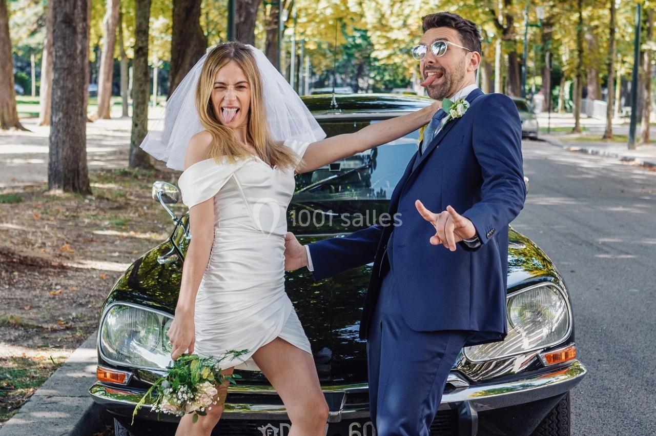 Un couple en tenue de mariage pose de manière décontractée et amusante devant une voiture ancienne dans un parc.