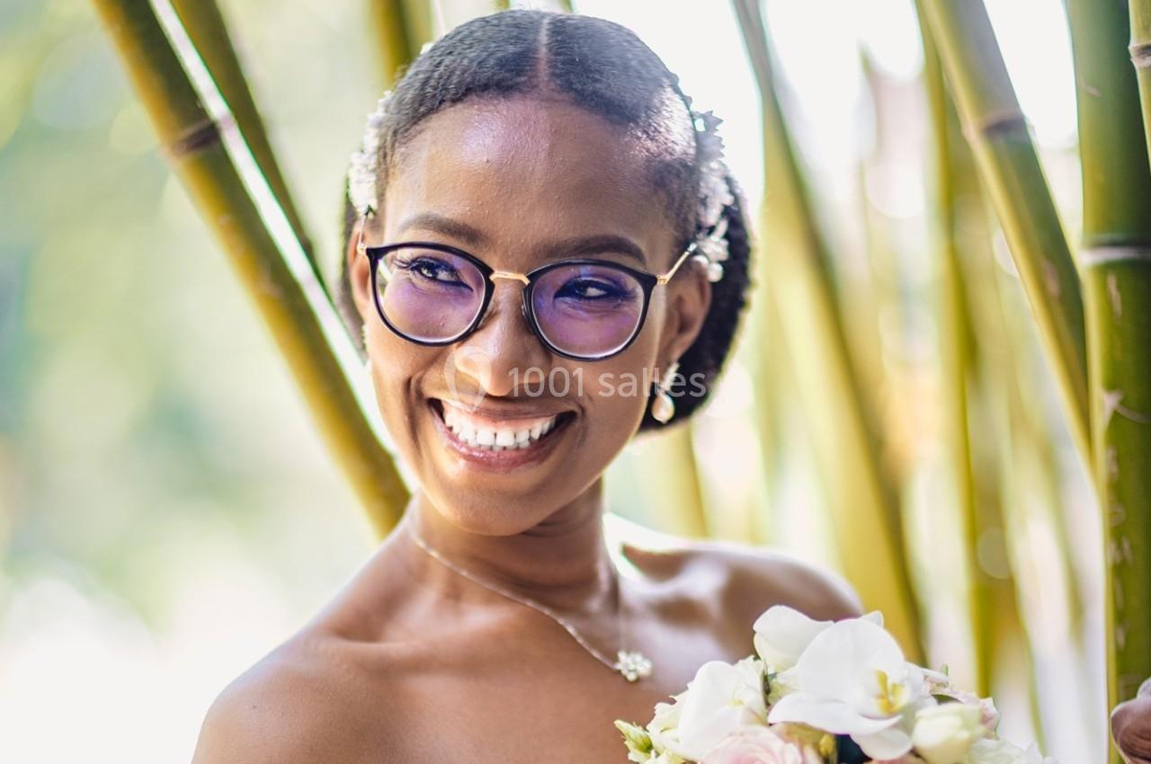 Une femme souriante portant des lunettes et tenant un bouquet de fleurs devant des bambous en arrière-plan flou.