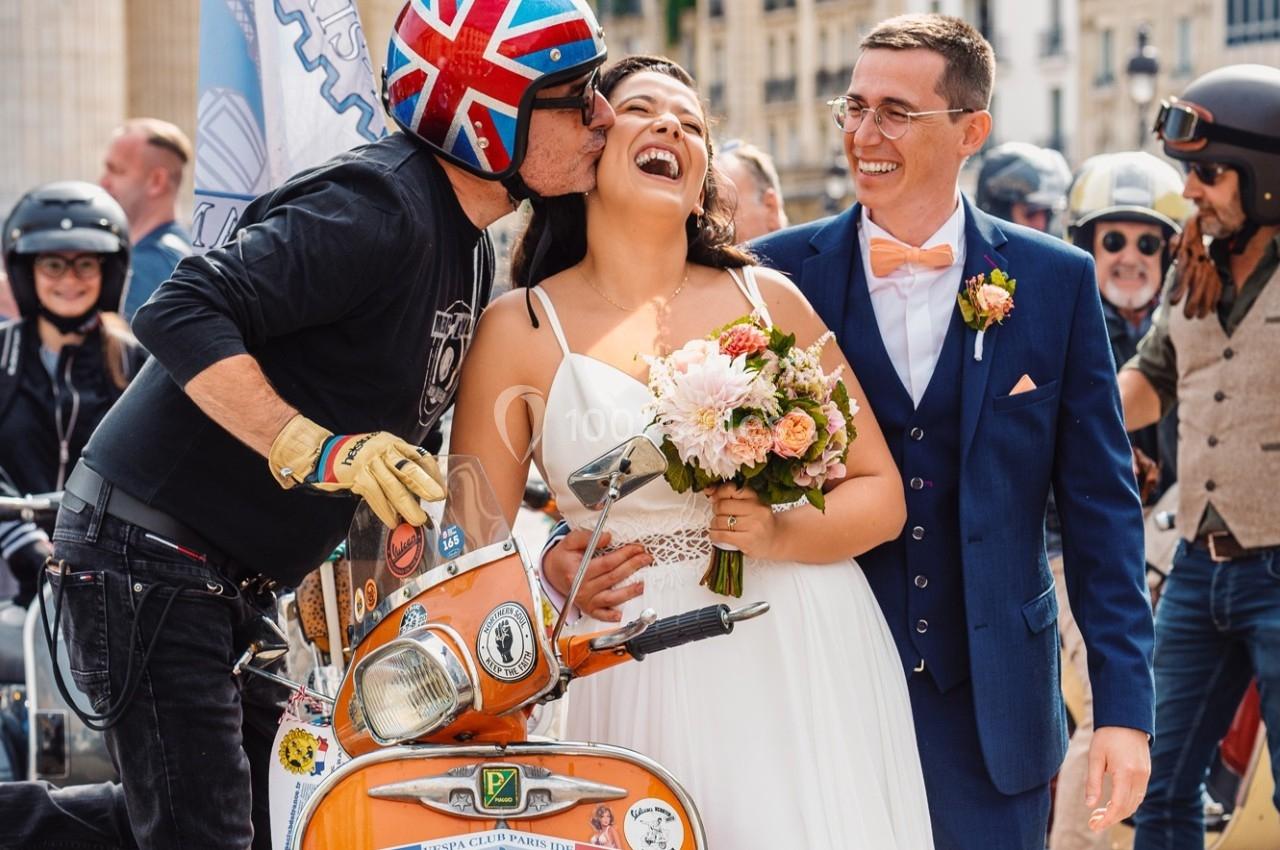 Un couple de mariés souriant, entouré d'un motard embrassant la mariée près d'un scooter orange.