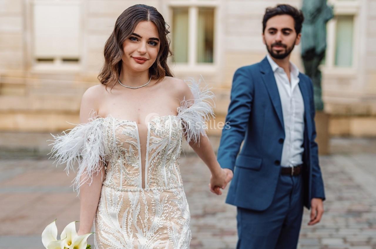 Une femme en robe de mariée tenant un bouquet, accompagnée d'un homme en costume dans un cadre urbain.