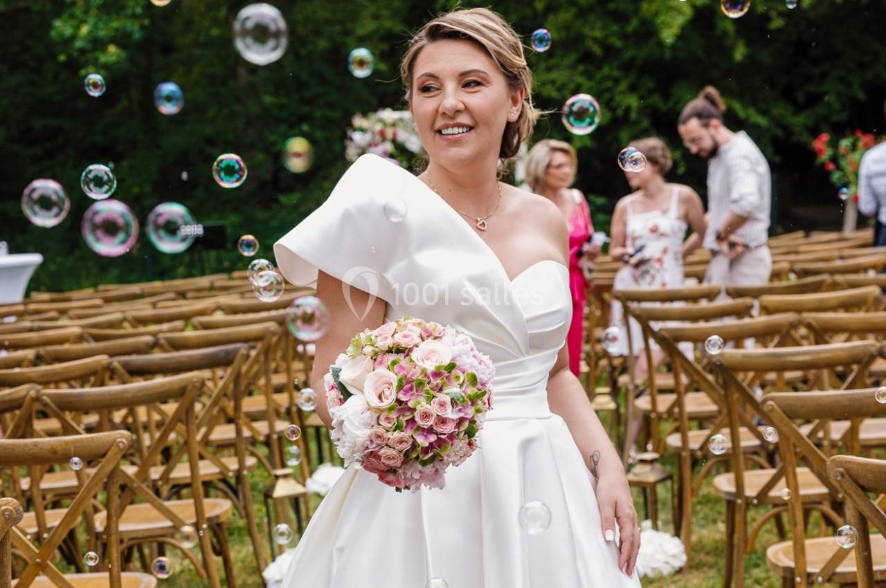Une mariée souriante en robe blanche tient un bouquet de fleurs roses, entourée de bulles et de chaises en bois.