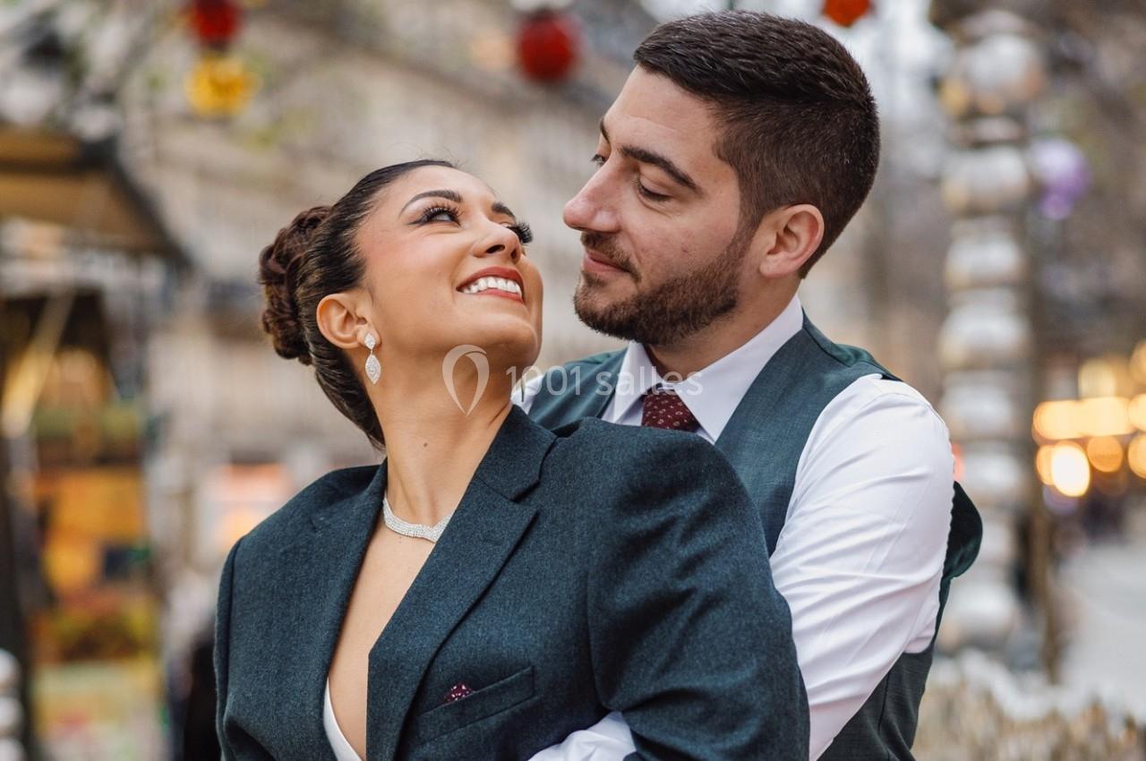 Un couple souriant s'enlace dans une rue décorée, avec un arrière-plan flou de lumières et de structures festives.