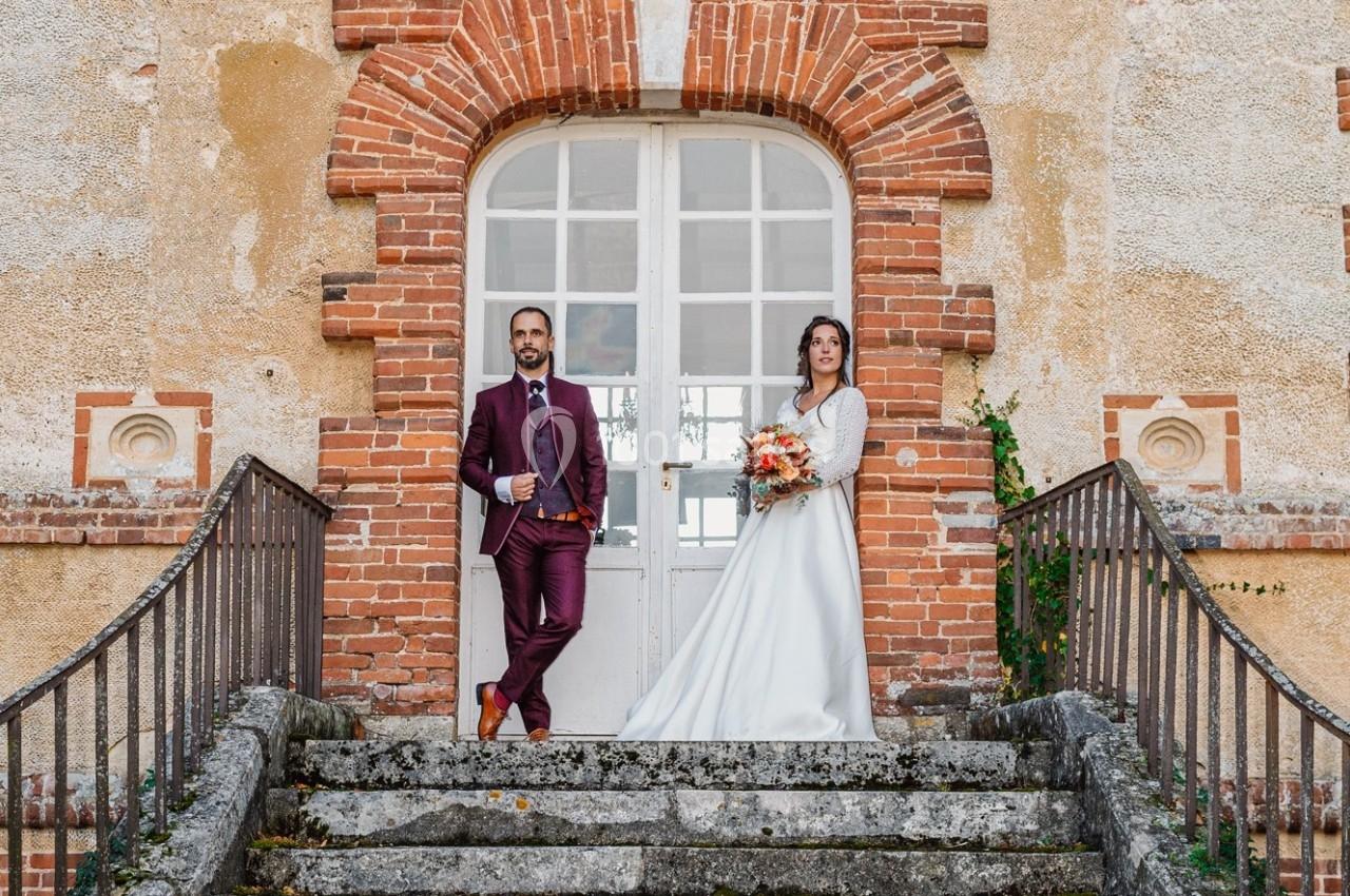 Un couple en tenue de mariage pose sur des marches devant une porte en arc entourée de briques.